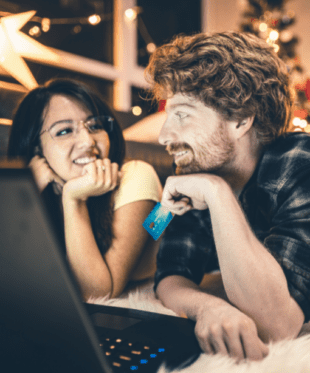 A man with a card in his hand and a woman with glasses are looking at each other in front of a laptop screen.
